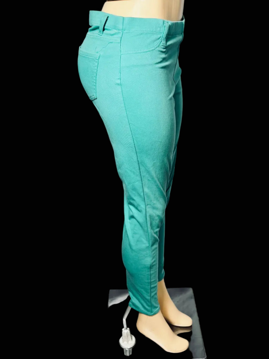 A pair of teal slim-fit pants displayed on a beige mannequin leg, standing on a small gray platform against a white draped backdrop.