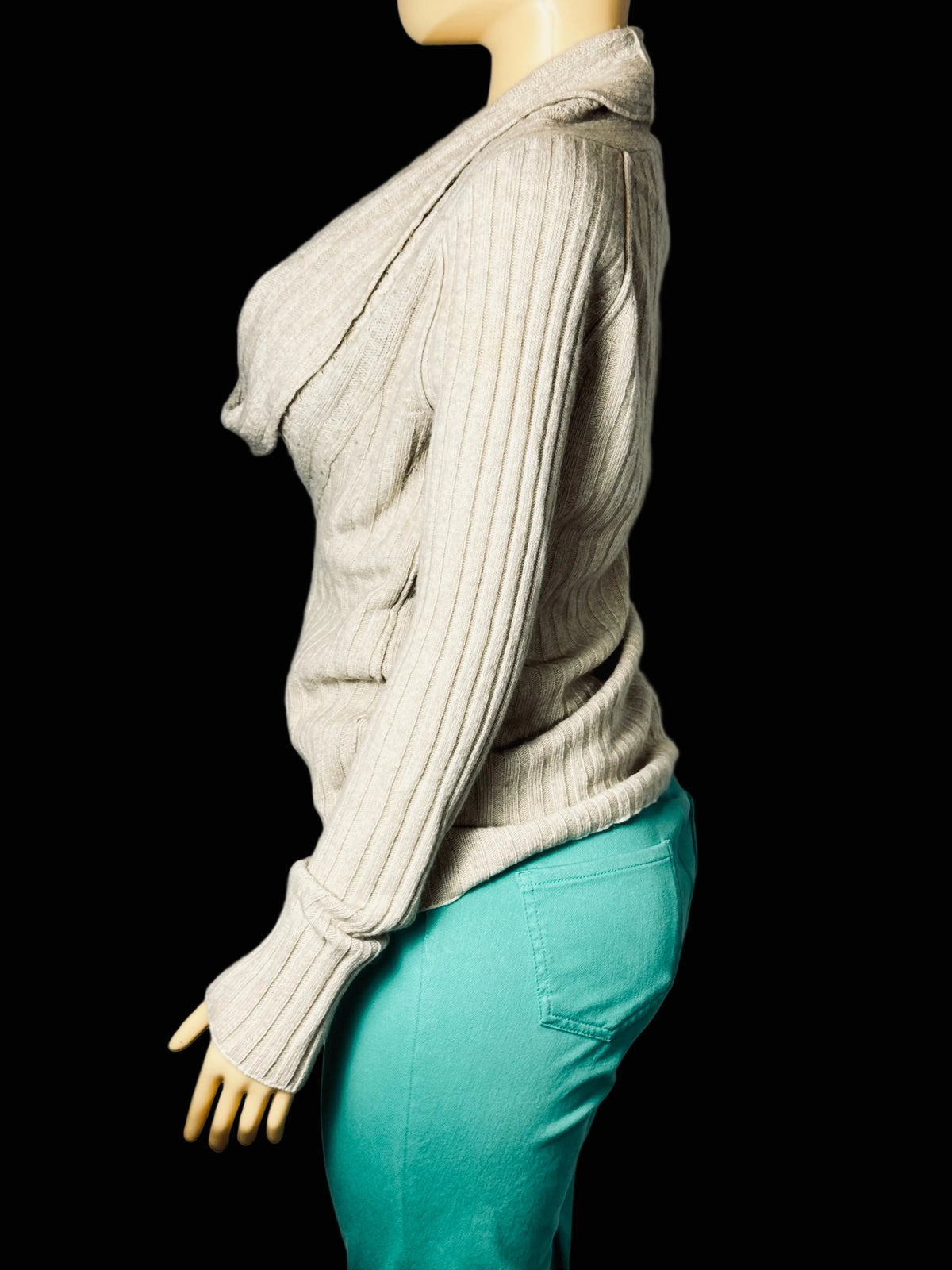 A side-profile view of a beige ribbed knit sweater with a cowl neck and thumbhole sleeves, displayed on a mannequin wearing teal pants against a light gray fabric backdrop.