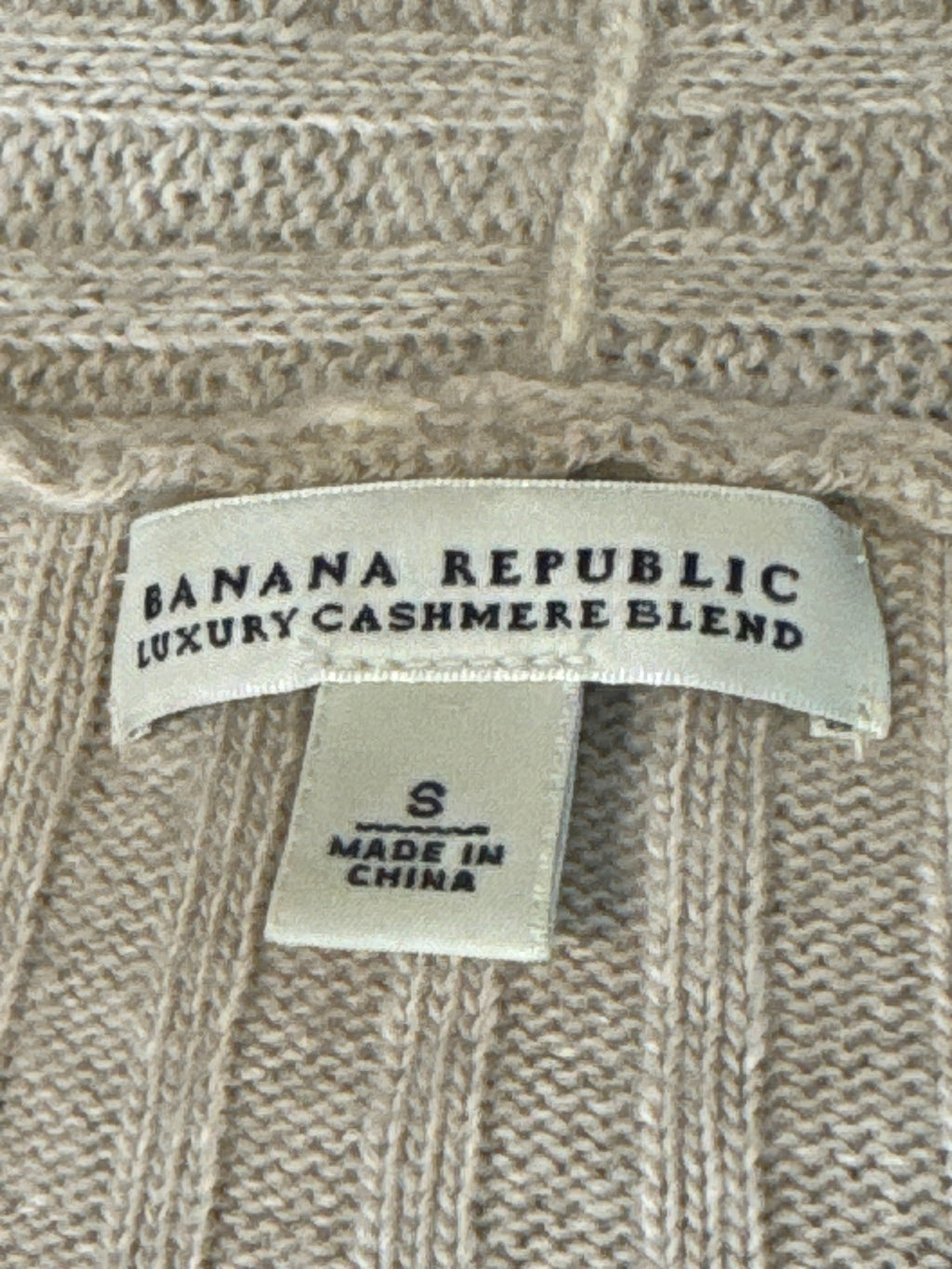 Close-up of a beige knit sweater label reading BANANA REPUBLIC LUXURY CASHMERE BLEND, with a smaller size tag below reading S and MADE IN CHINA.