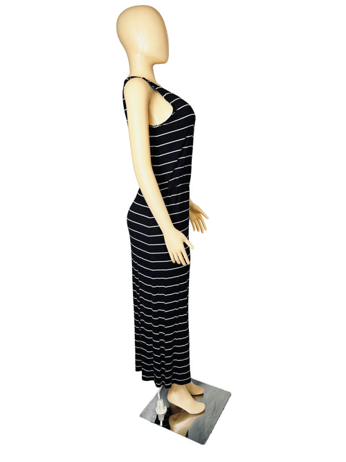 A sleeveless black maxi dress with thin white horizontal stripes displayed on a beige mannequin. The mannequin is standing on a small mirror on a carpeted floor in front of a white fabric backdrop, with a wooden dresser visible on the left.