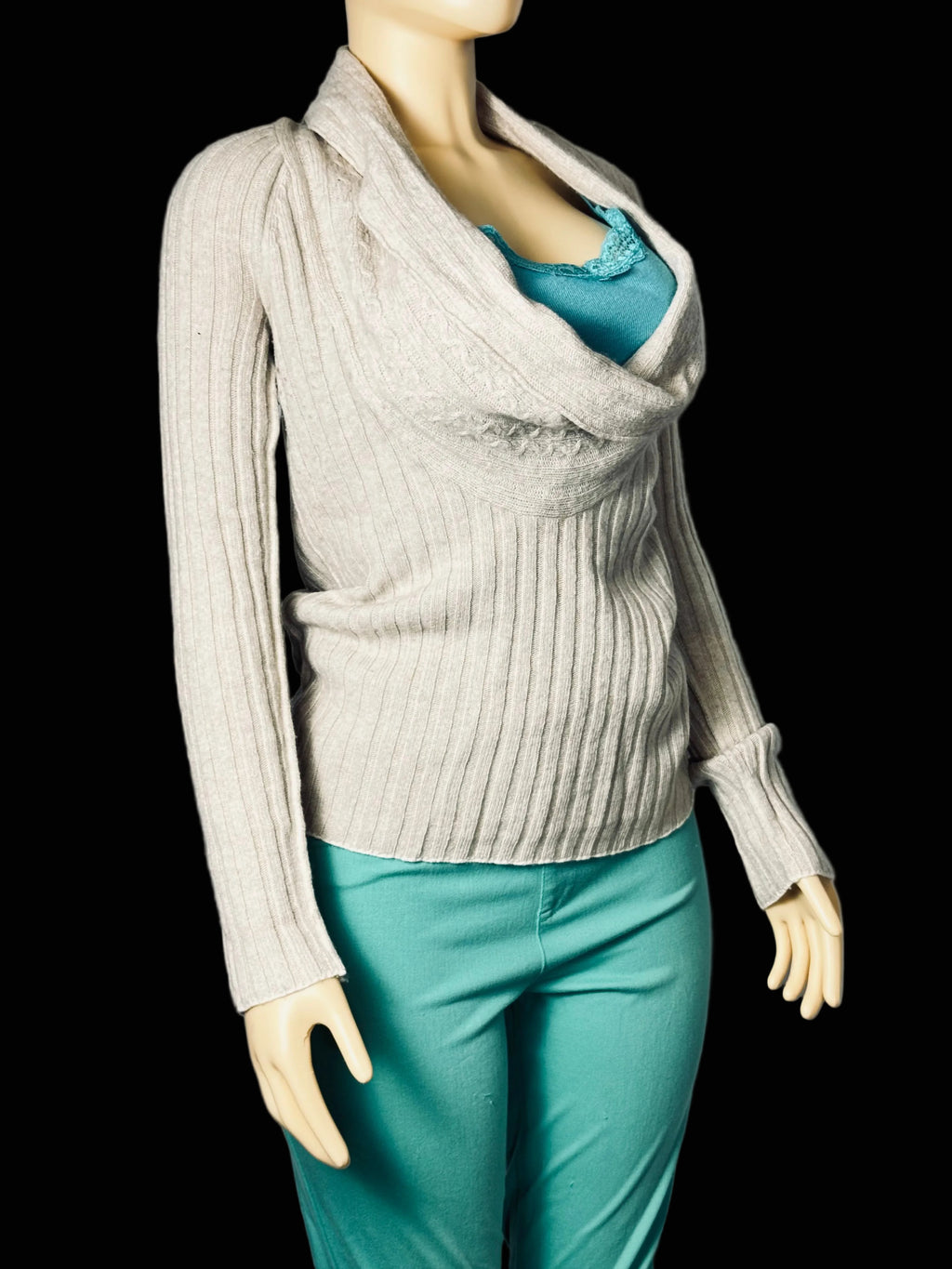A ribbed knit cowl-neck sweater in light beige, displayed on a mannequin torso with a teal top and teal pants against a gray fabric backdrop.