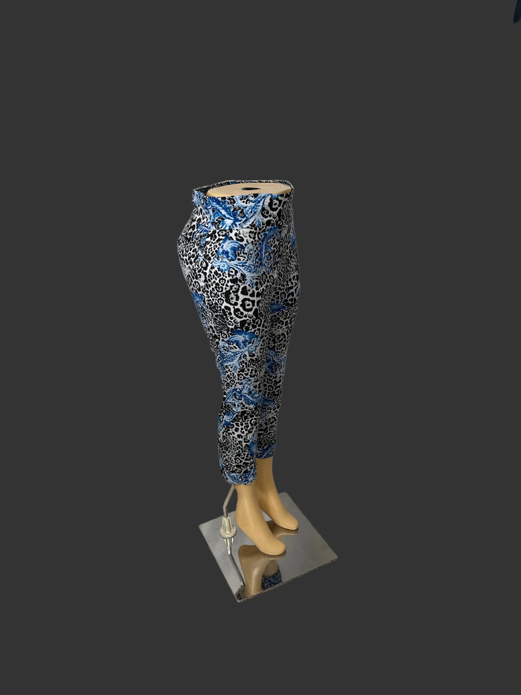 Patterned ankle-length pants displayed on a mannequin lower body with beige feet, set against a white draped fabric backdrop in an indoor room with carpet and clothing visible at the edges.