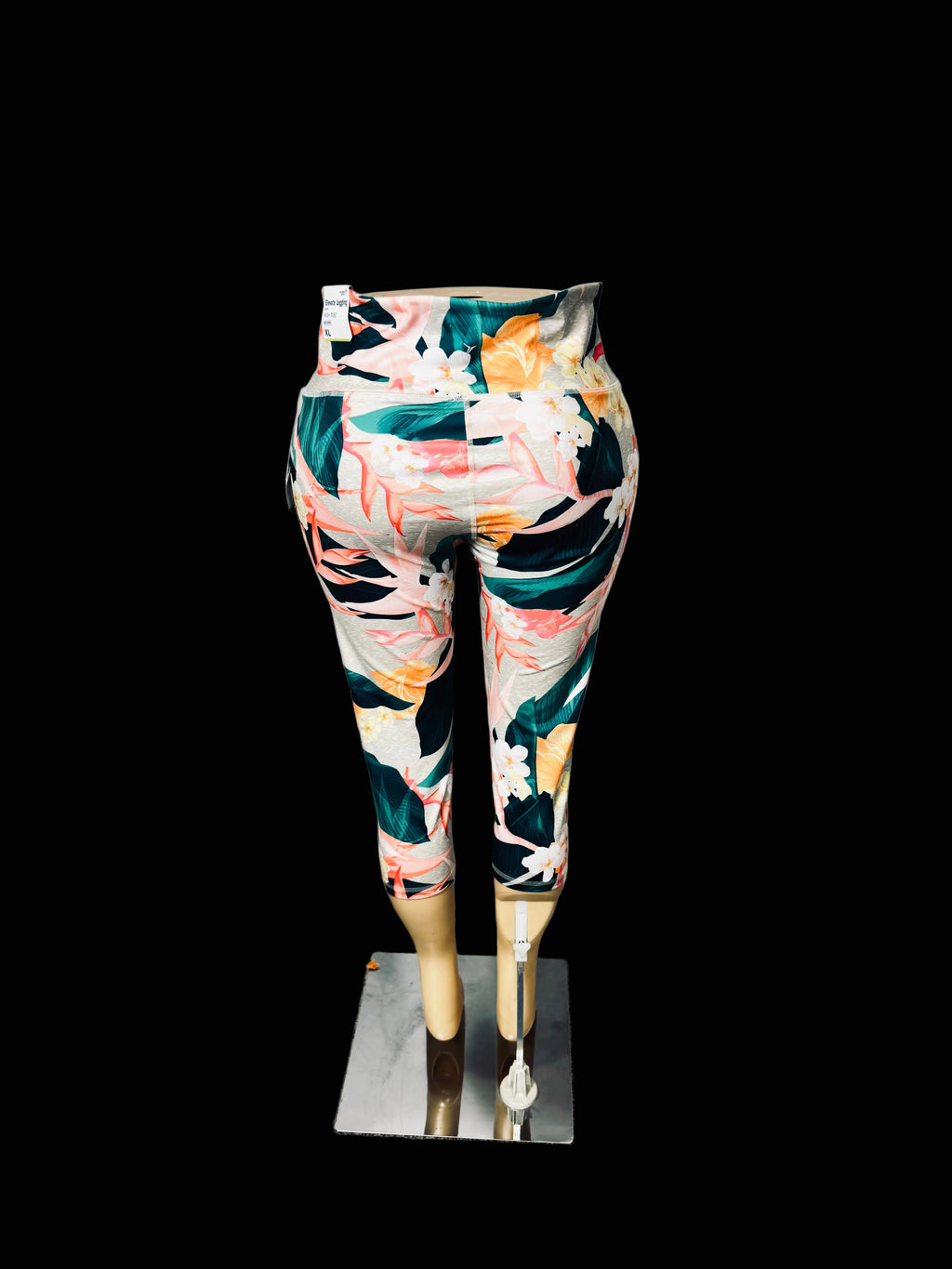 High-waisted capri leggings with a colorful abstract botanical print in teal, orange, pink, black, and white, displayed on a mannequin from the back against a white draped backdrop.