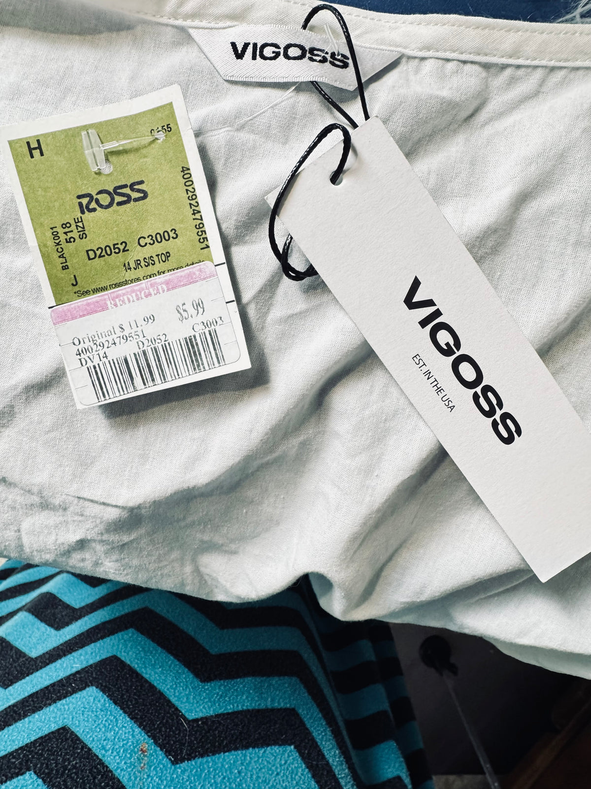 A close-up of a light gray or off-white VIGOSS garment laid flat, with a large white retail hangtag and a smaller size/price tag attached. The VIGOSS brand name is clearly visible on both the neck label and the hangtag.