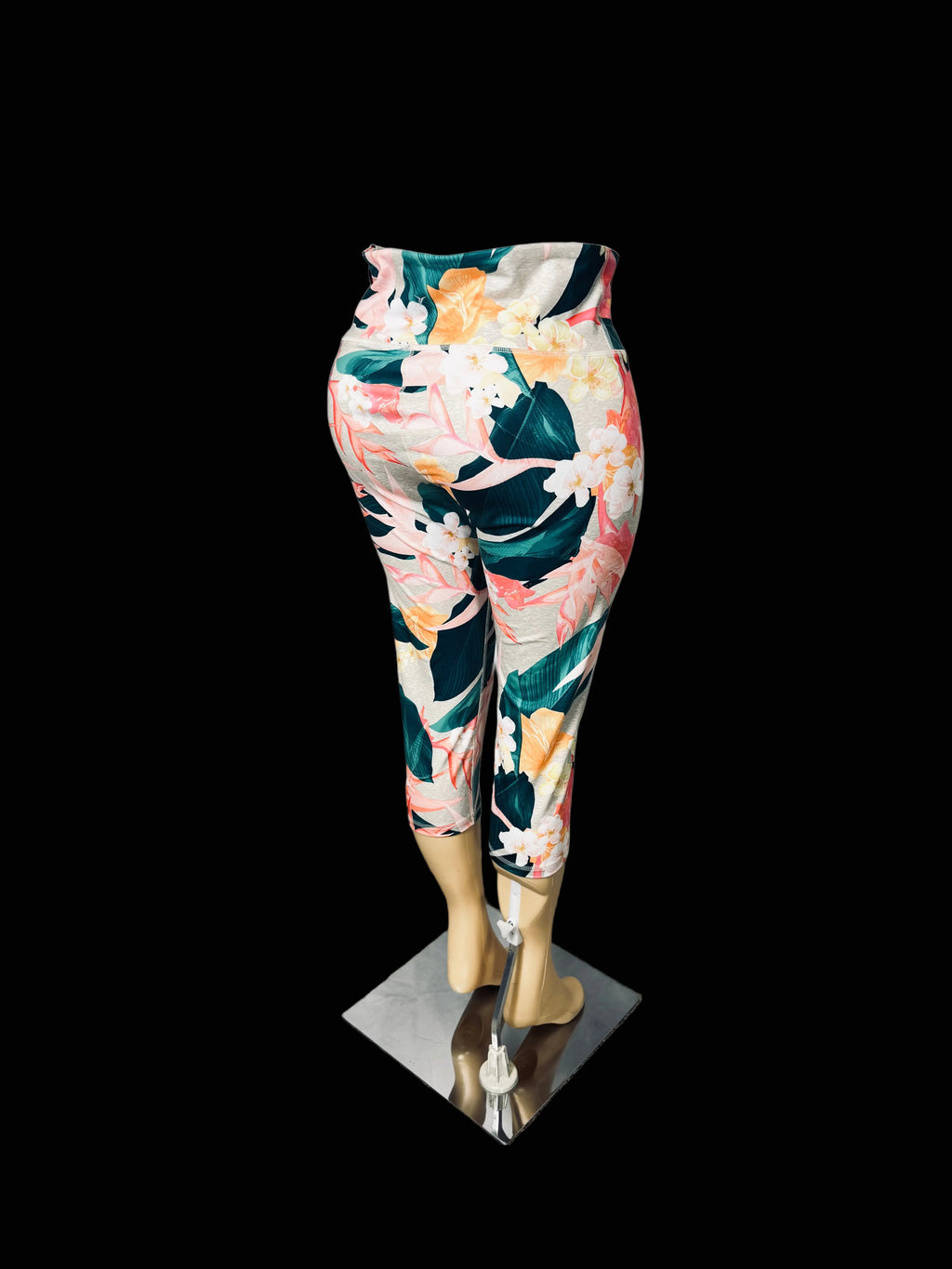 A pair of high-waisted, cropped leggings with a vibrant tropical floral print, displayed on a mannequin leg form against a white fabric backdrop.
