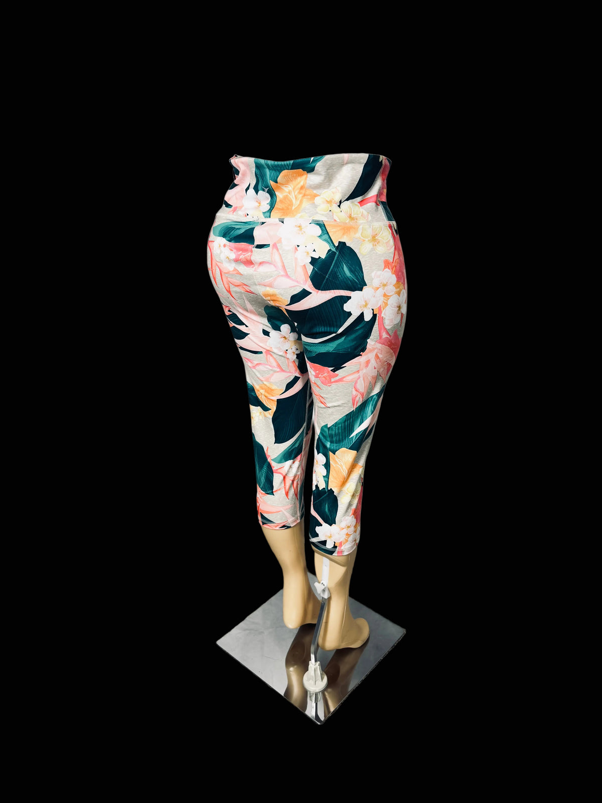A pair of high-waisted, cropped leggings with a vibrant tropical floral print, displayed on a mannequin leg form against a white fabric backdrop.