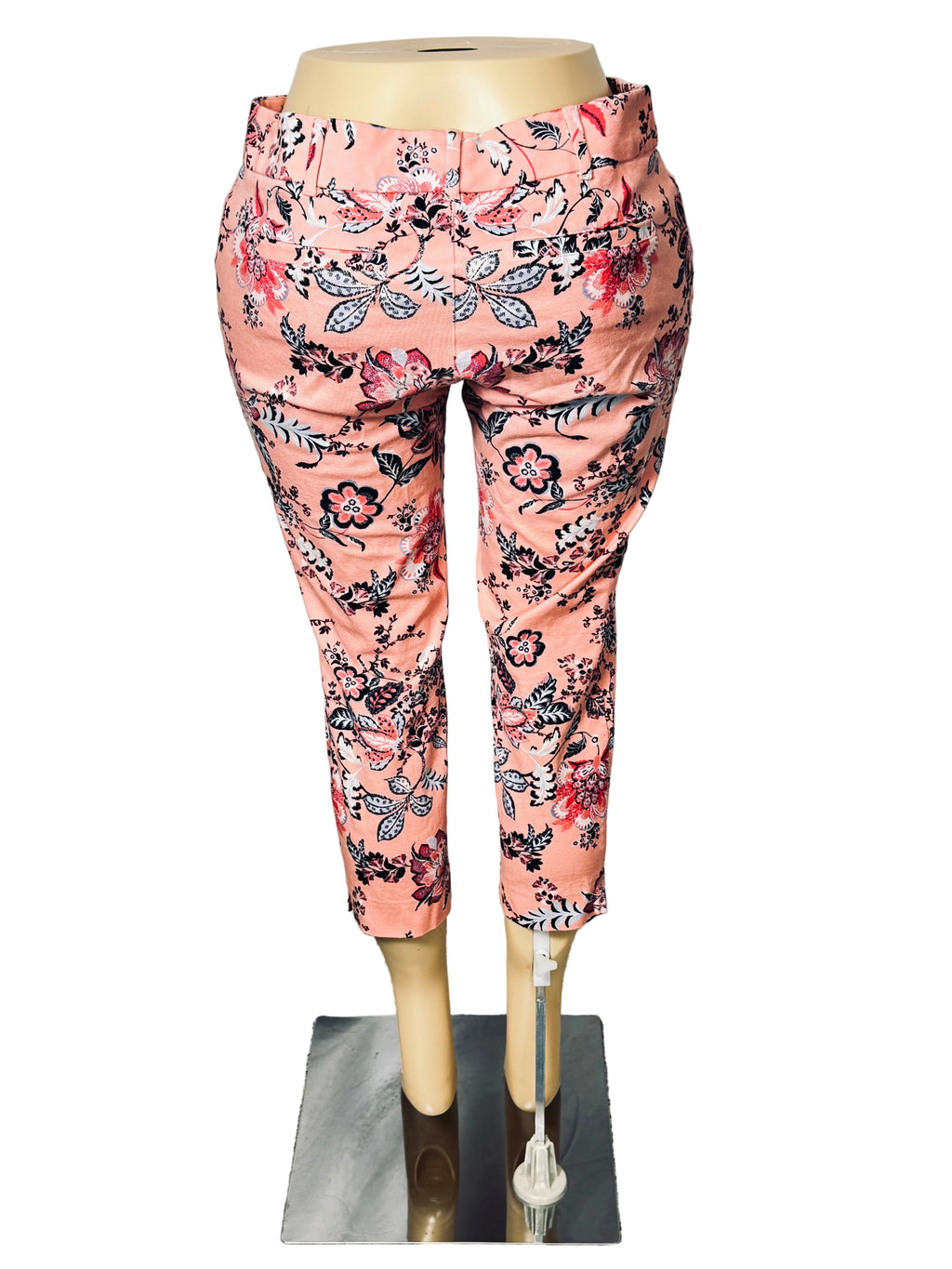 Floral print cropped pants in a pinkish-orange tone with black, red, and blue botanical patterns, displayed on a mannequin against a white draped backdrop.