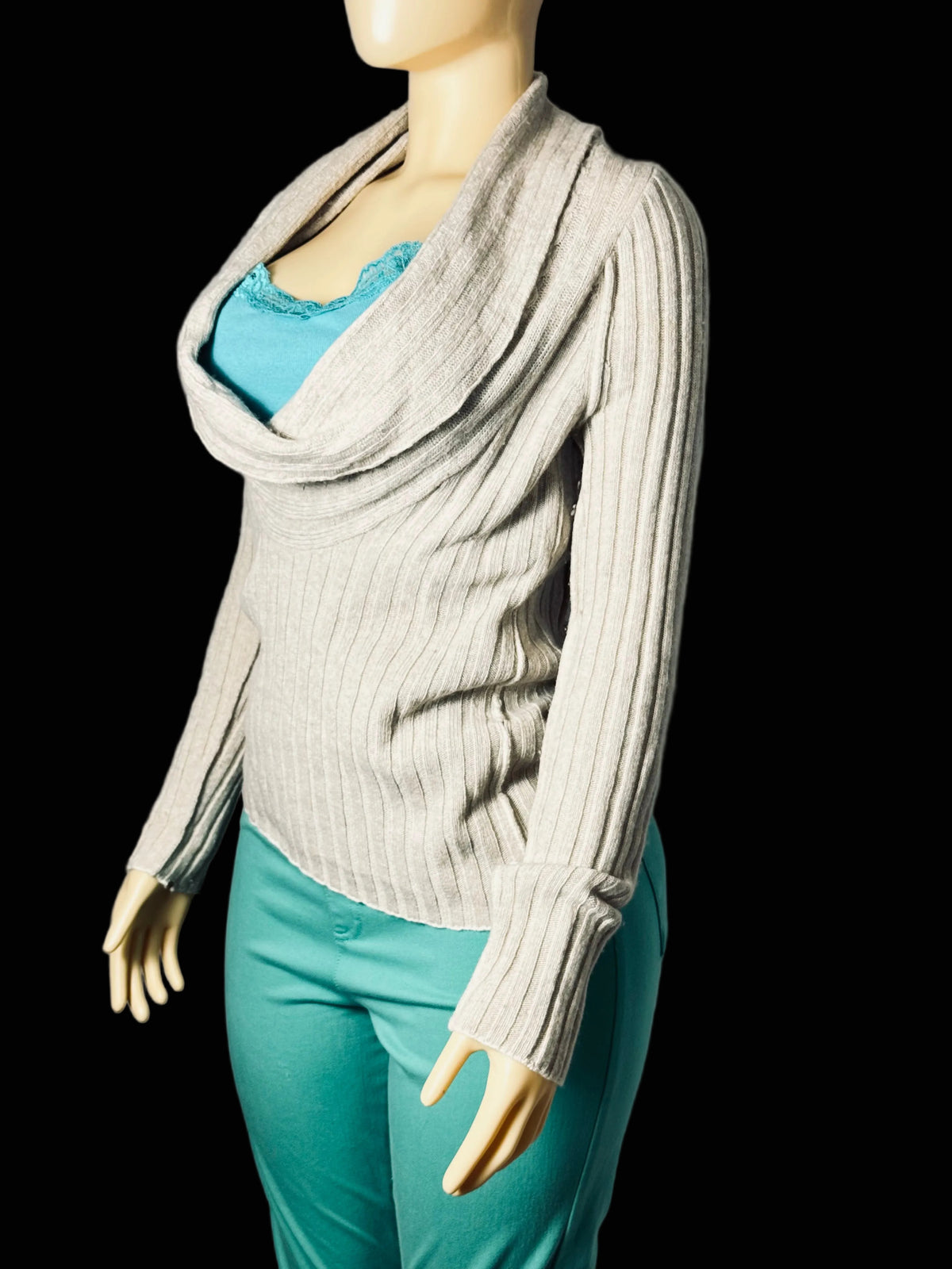 A mannequin wearing a ribbed knit cowl-neck sweater in a light taupe-beige color, layered over a turquoise top and paired with turquoise pants, set against a wrinkled gray fabric backdrop.